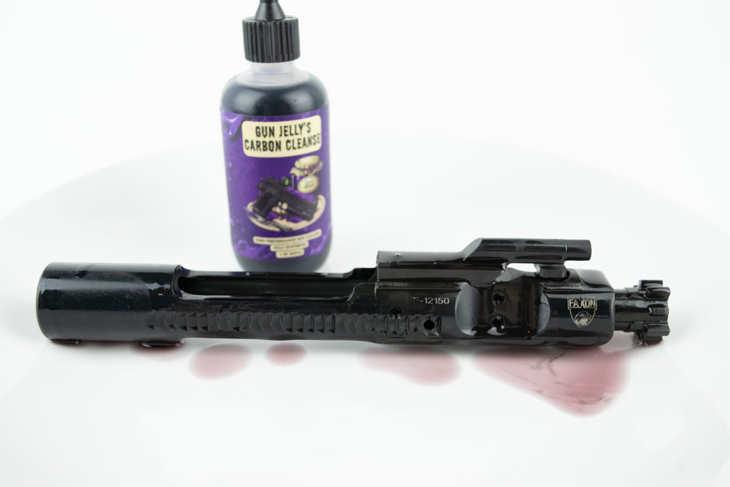 A black bolt carrier group lies horizontally on a white surface with a small puddle of purple liquid in front of it. A purple bottle labeled 'Gun Jelly's Carbon Cleanse' stands behind it on a white studio background.
