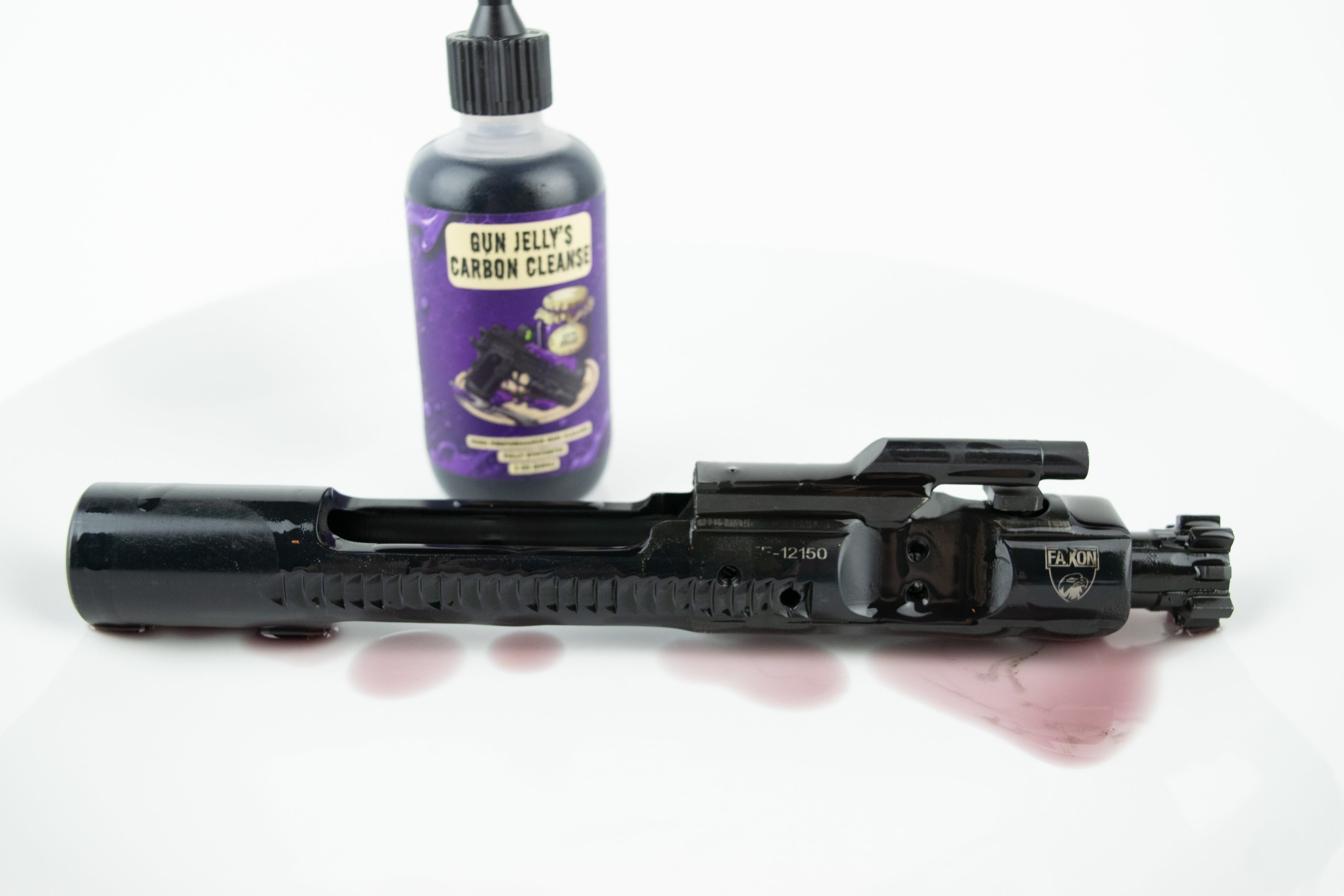 A black bolt carrier group lies horizontally on a white surface with a small puddle of purple liquid in front of it. A purple bottle labeled 'Gun Jelly's Carbon Cleanse' stands behind it on a white studio background.