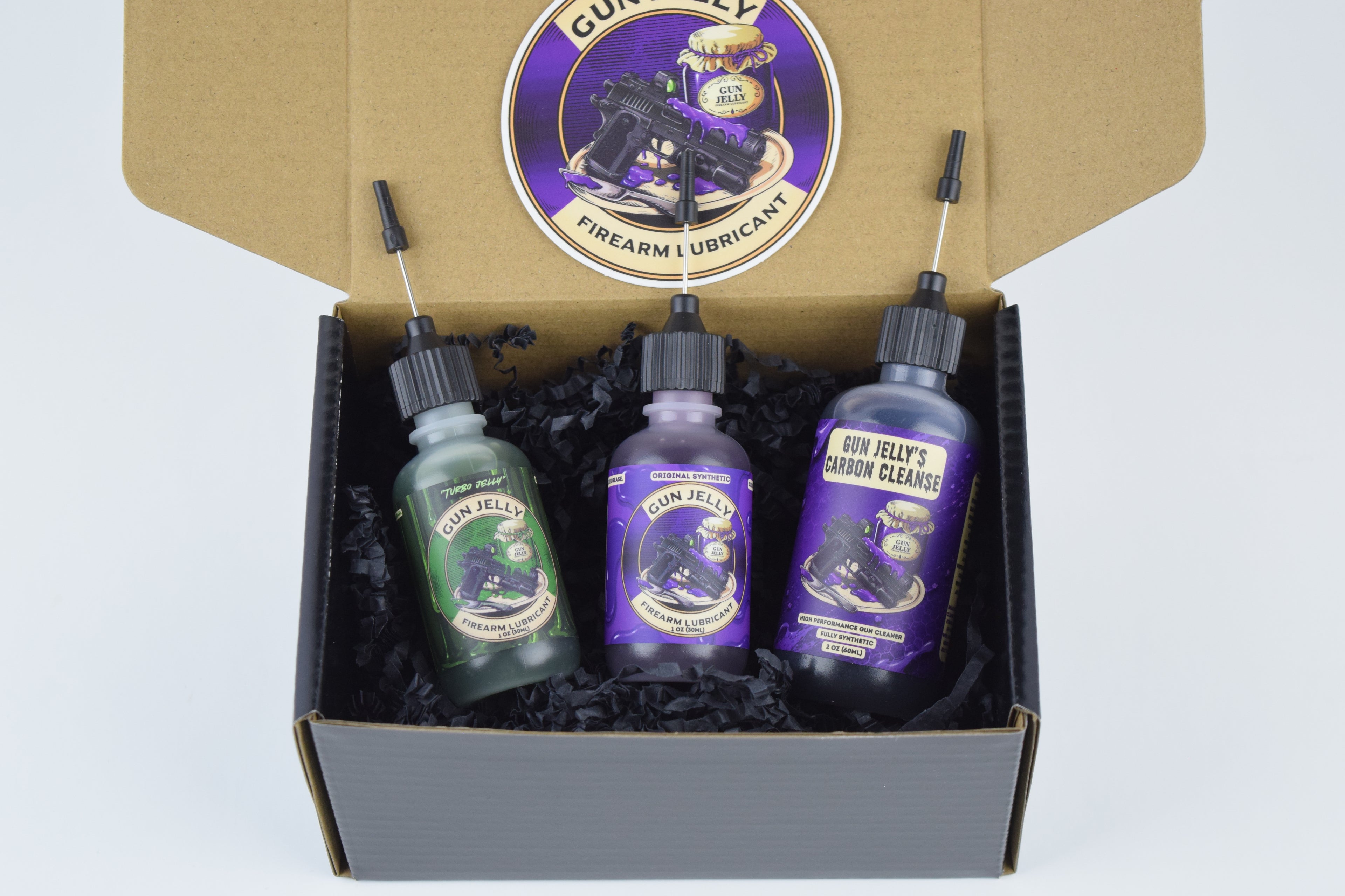 A gun care gift set in a brown cardboard box with black crinkle paper filler, featuring three bottles of Gun Jelly lubricant and a bottle of Gun Jelly Carbon Cleanse Cleaner, with the brand logo visible on the inside lid as a sticker.